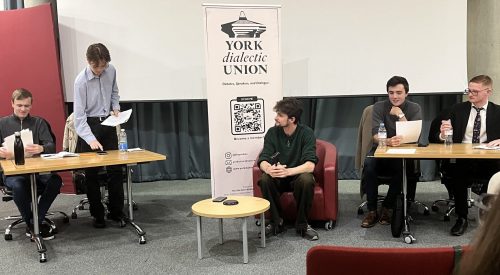 Four speakers and a judge sitting at two panel tables, preparing to speak.