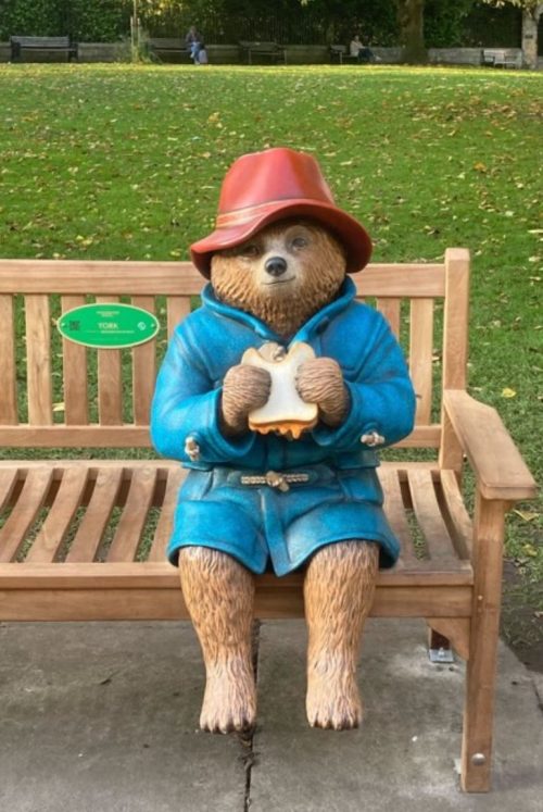 Paddington bear sitting down on a bench in a blue overcoat wearing a red hat eating a sandwich