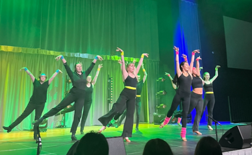 A dozen Ballroom and Latin dancers in all black on stage with green lights behind them