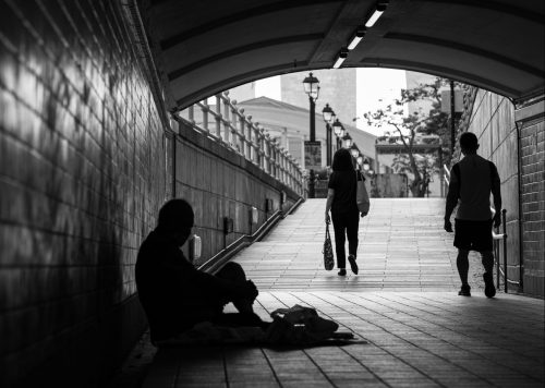 Black and white photo where a homeless man sits under a bridge surrounded by blankets as passers-by walk past him with shopping bags