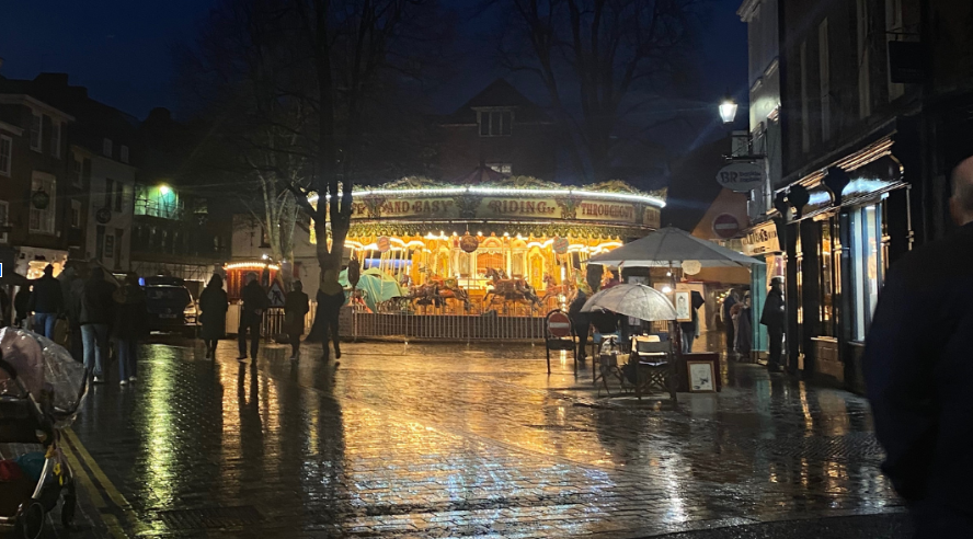 Centrally, a brightly lit carousel shines in the centre of a dark cobbled street, a few people in warm clothes walk either side.