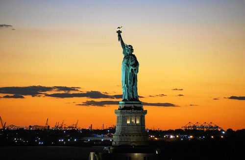 The Statue of Liberty is central, with an orange sunset in the background. Lights from dark buildings in distance can be seen at the bottom of the screen.