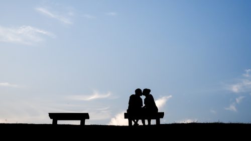 A silhouette of a couple kiss each other on a bench in front of a blue sky.