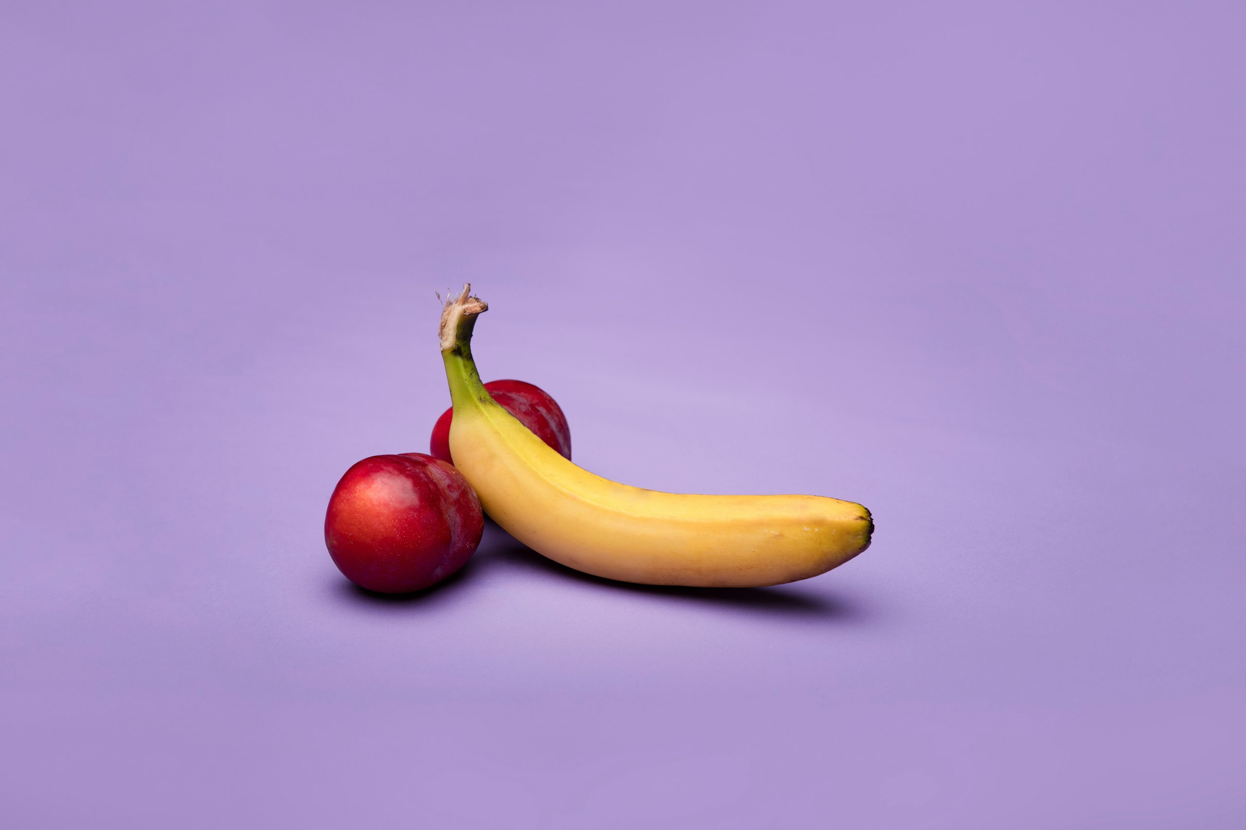 Two red plums and a yellow banana in front of a purple background.